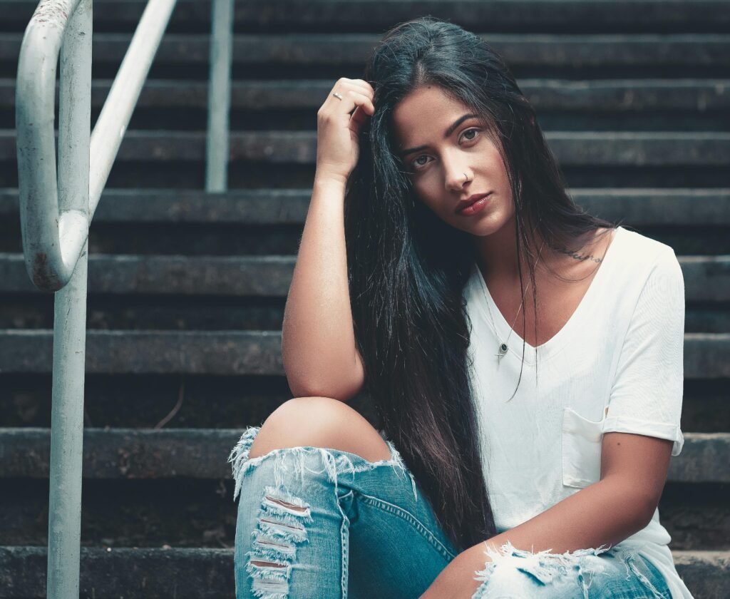 Portrait of a young woman with long dark hair sitting on stairs in casual fashion attire.